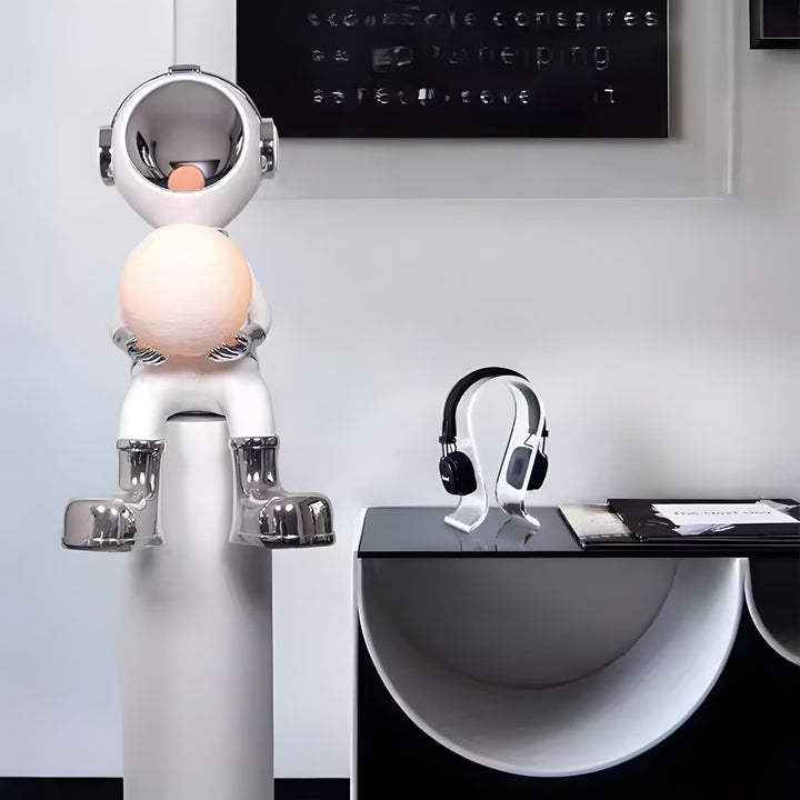 Sculptural astronaut lamp holding a glowing orb next to stylish headphones on a modern table.