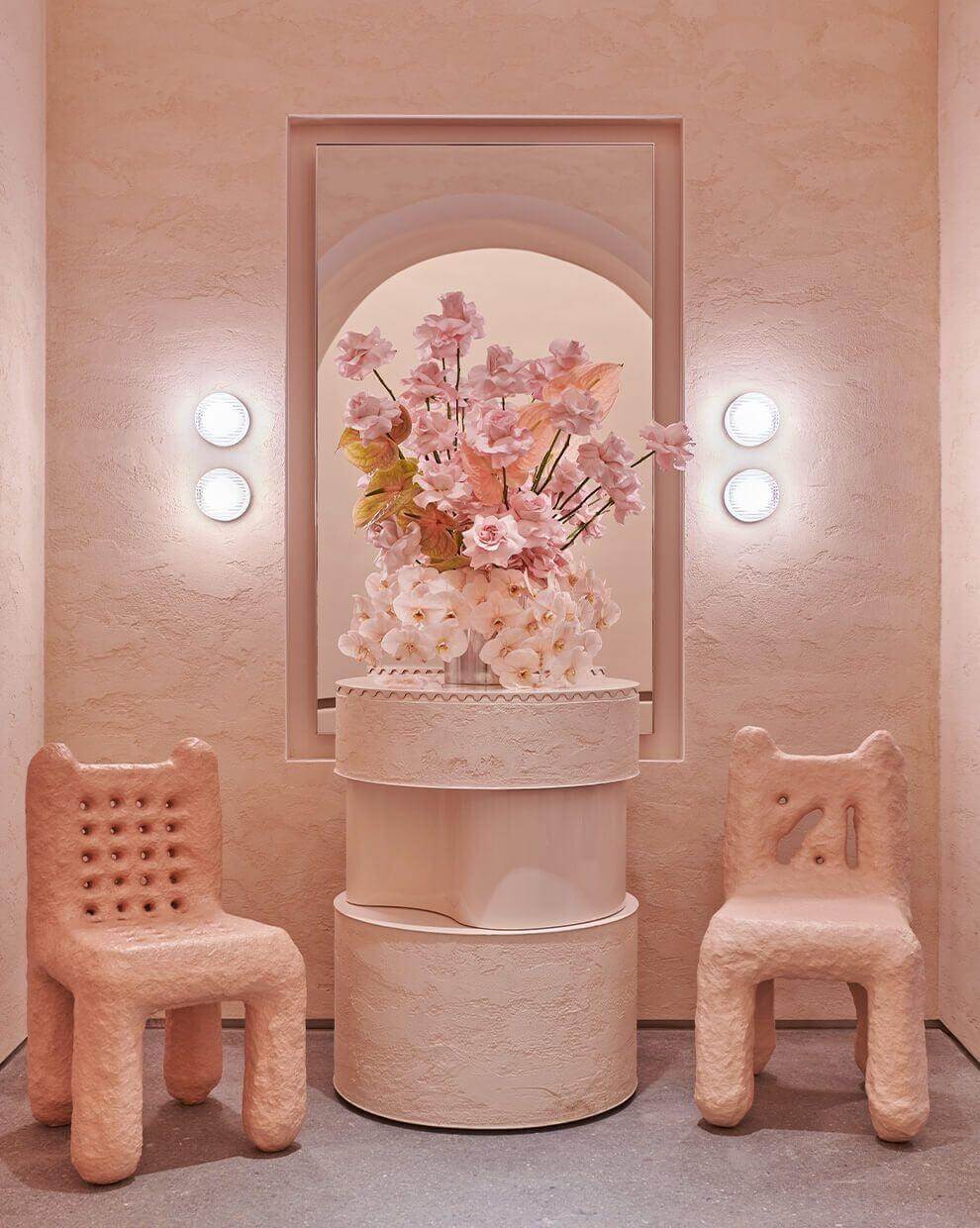 Interior design featuring pink decor, two unique chairs, a floral arrangement, and modern wall sconces.