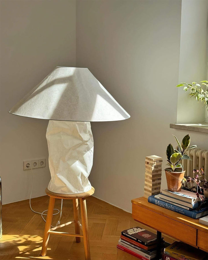 Minimal white table lamp on a stool, casting shadows in a cozy bedroom setting with books and plants.