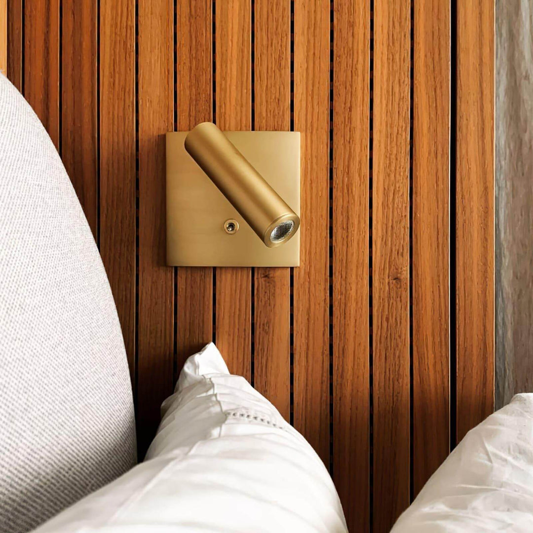 Micro Square Switched Sconce in gold finish mounted on wooden wall beside bed, ideal for minimalist bedside lighting.
