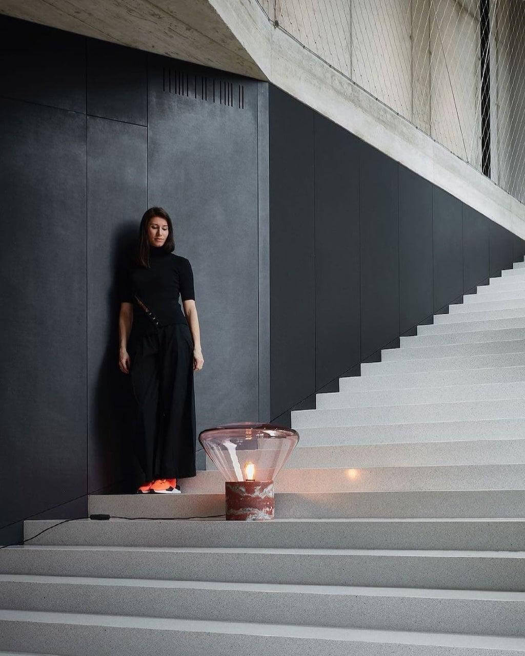 Elegant woman in black outfit beside a stylish round glass table lamp on minimalist stairs, showcasing modern lighting design.
