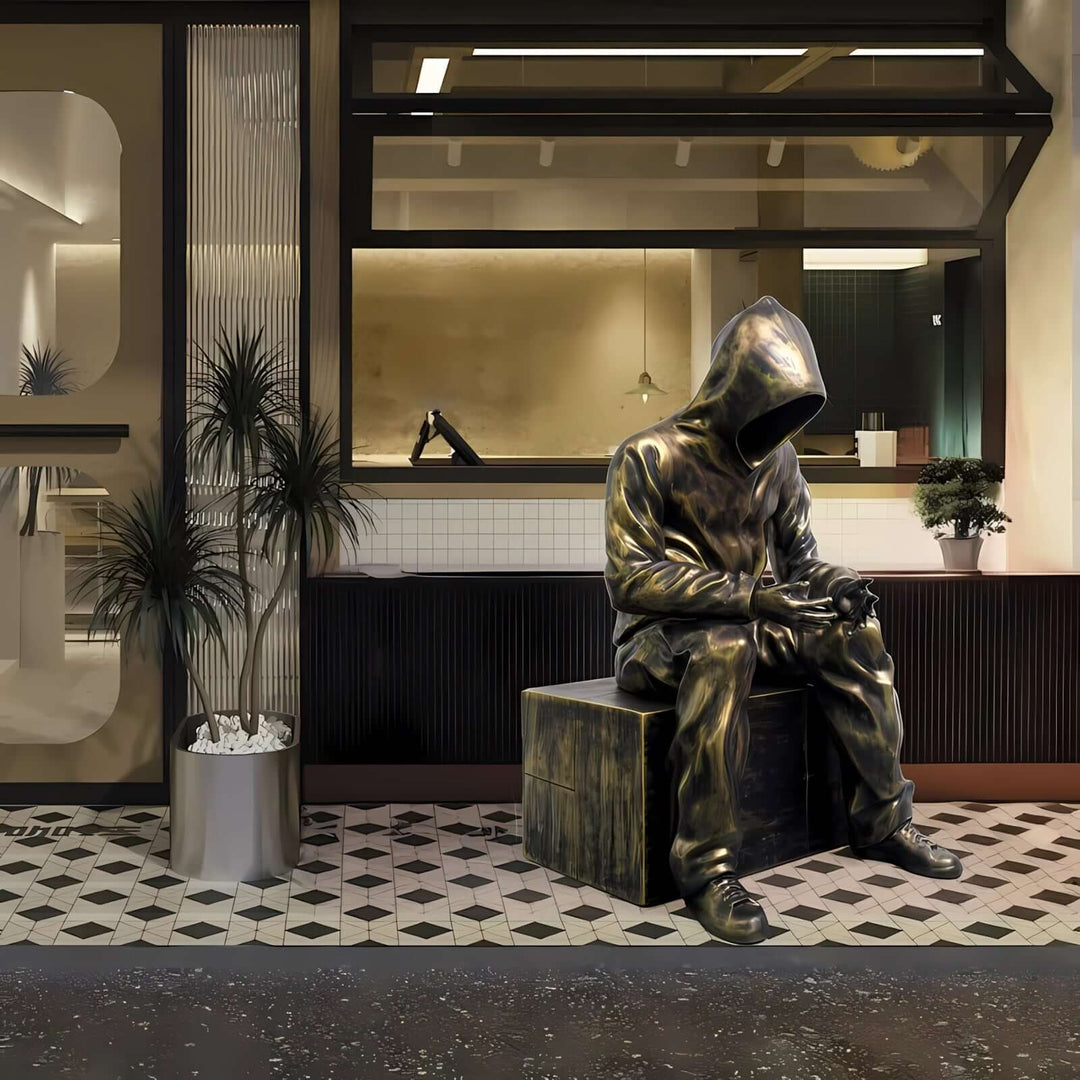 Sculptural bronze statue of a hooded figure seated on a block in a stylish café setting.