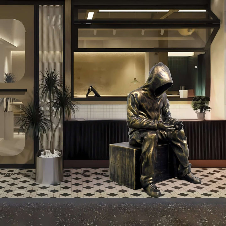 Sculptural bronze statue of a hooded figure seated on a block in a stylish café setting.