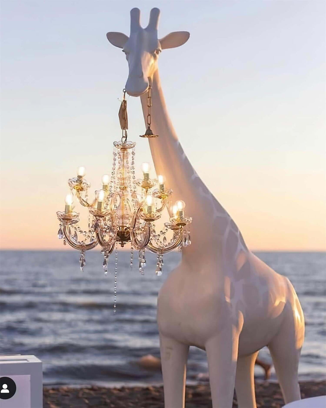 Stylish sculptural giraffe floor lamp holding a chandelier by the beach at sunset, perfect for modern decor.
