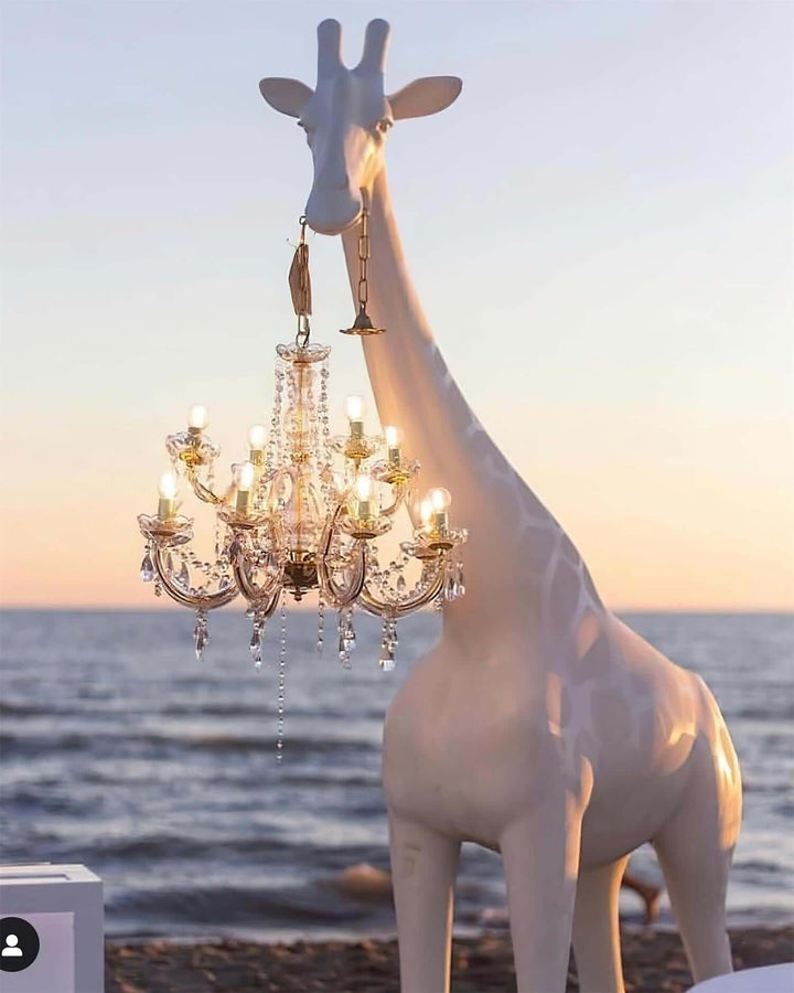 Stylish sculptural giraffe floor lamp holding a chandelier by the beach at sunset, perfect for modern decor.