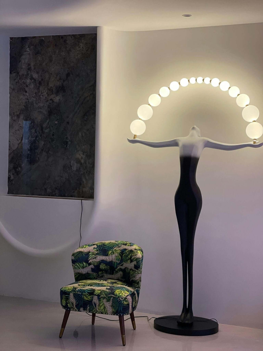 Sculptural floor lamp with a black figure base and arch of white bulbs, beside a colorful patterned chair in modern interior.