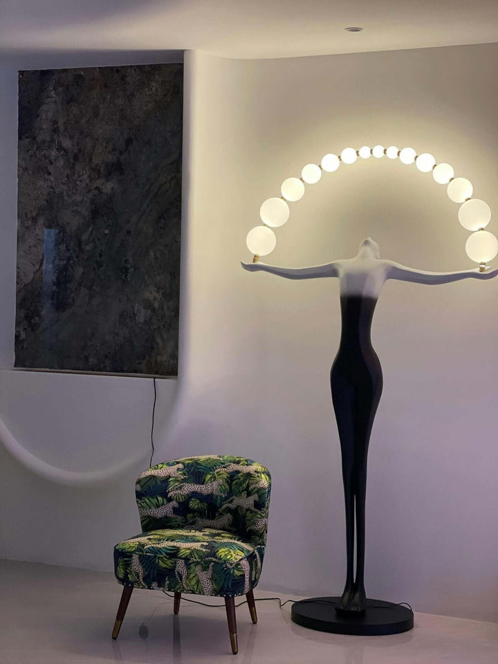 Sculptural floor lamp with a black figure base and arch of white bulbs, beside a colorful patterned chair in modern interior.