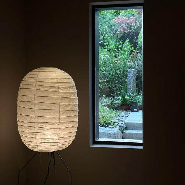 Decorative white washi paper floor lamp beside a window showcasing a serene outdoor garden view.