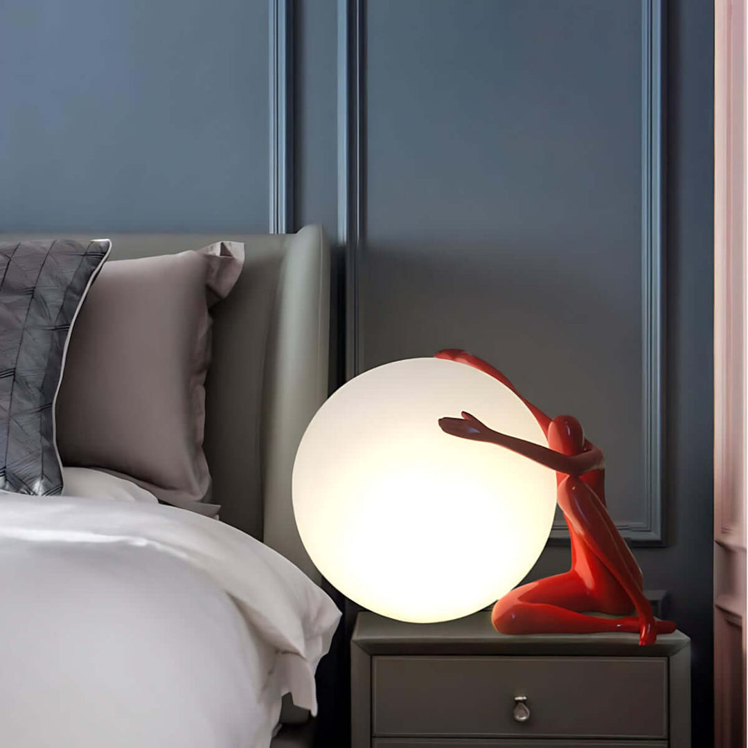 Zelvora sculptural table lamp beside a bed, featuring a red figure supporting a glowing white orb, ideal for modern decor.