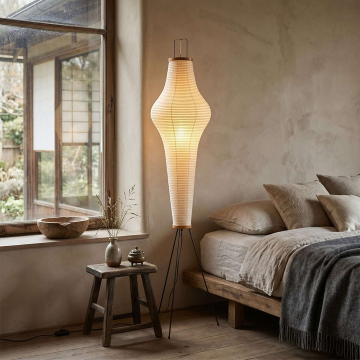 Akari 14A paper floor lamp glowing beside a wood platform bed in a warm, rustic bedroom.