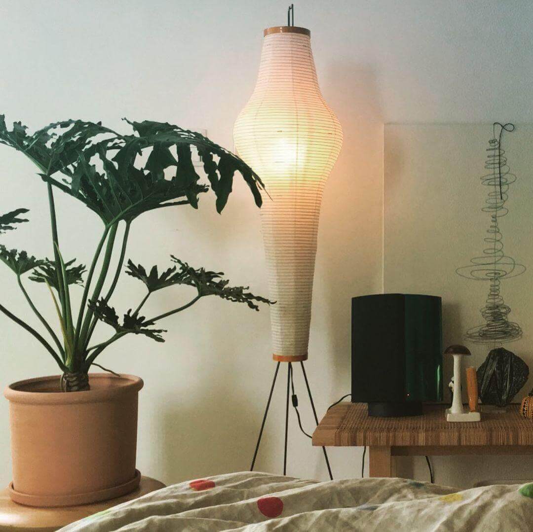 Akari 14A paper floor lamp glowing beside a large potted plant and wood console in a relaxed bedroom.