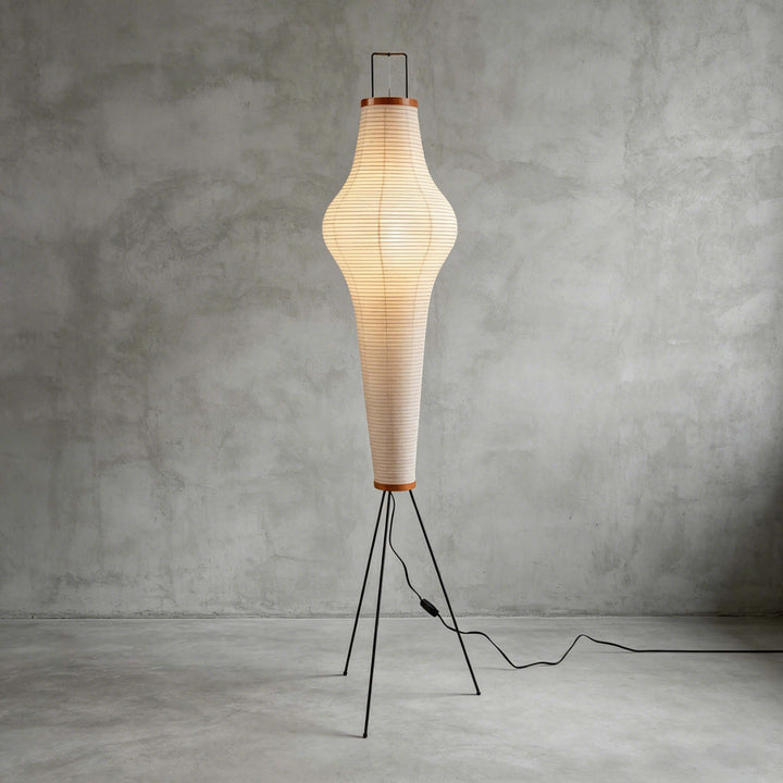 Akari 14A white washi paper floor lamp glowing against a gray concrete wall in a minimalist studio setting.