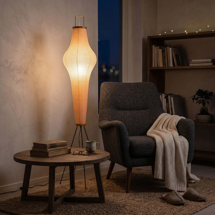 Akari 14A floor lamp casting a warm glow beside a gray armchair and bookshelf in an evening reading nook.