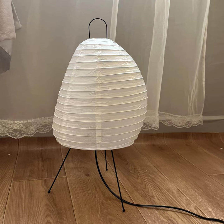 Unlit Akari 1N rice paper table lamp on a wood floor beside sheer curtains