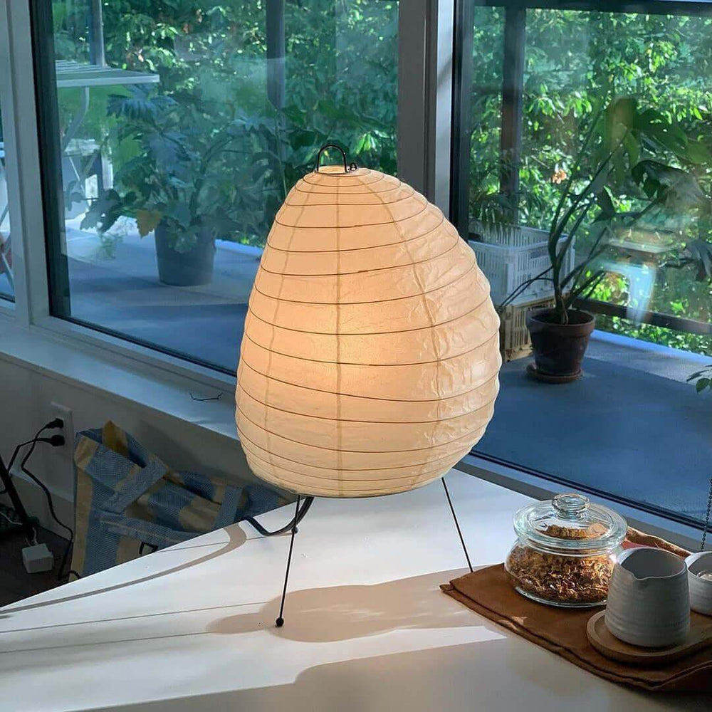 Glowing Akari 1N rice paper table lamp on a white table beside a leafy window