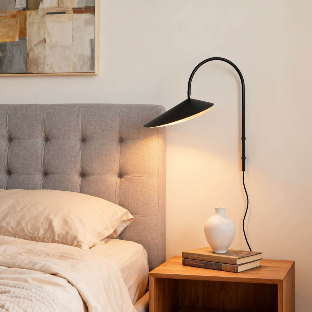 Black Arum plug-in wall sconce casting warm light beside a gray upholstered bed and wood nightstand