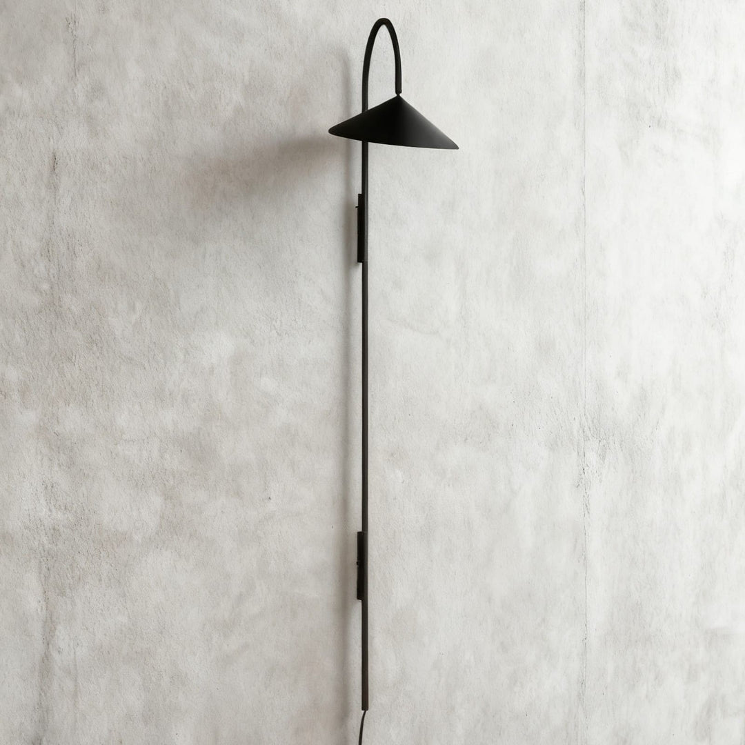 Long black Arum plug-in wall sconce with an extended vertical stem displayed on a gray plaster wall