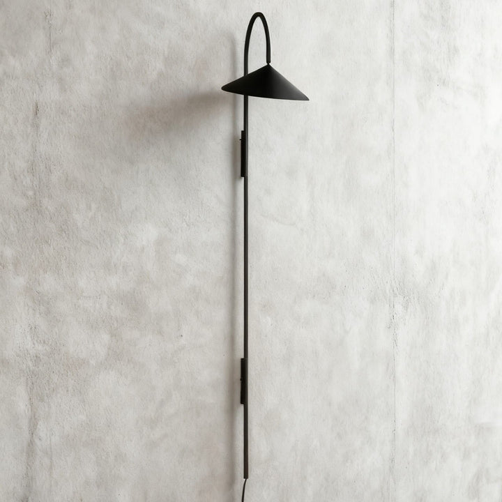 Long black Arum plug-in wall sconce with an extended vertical stem displayed on a gray plaster wall