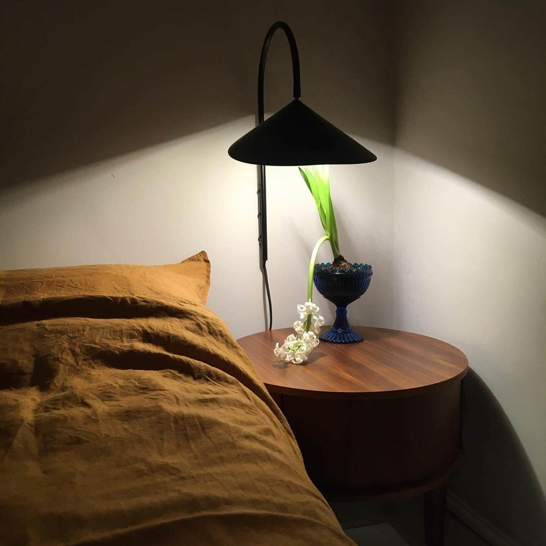 Black Arum wall-mounted reading light above a round wood bedside table in a dark bedroom