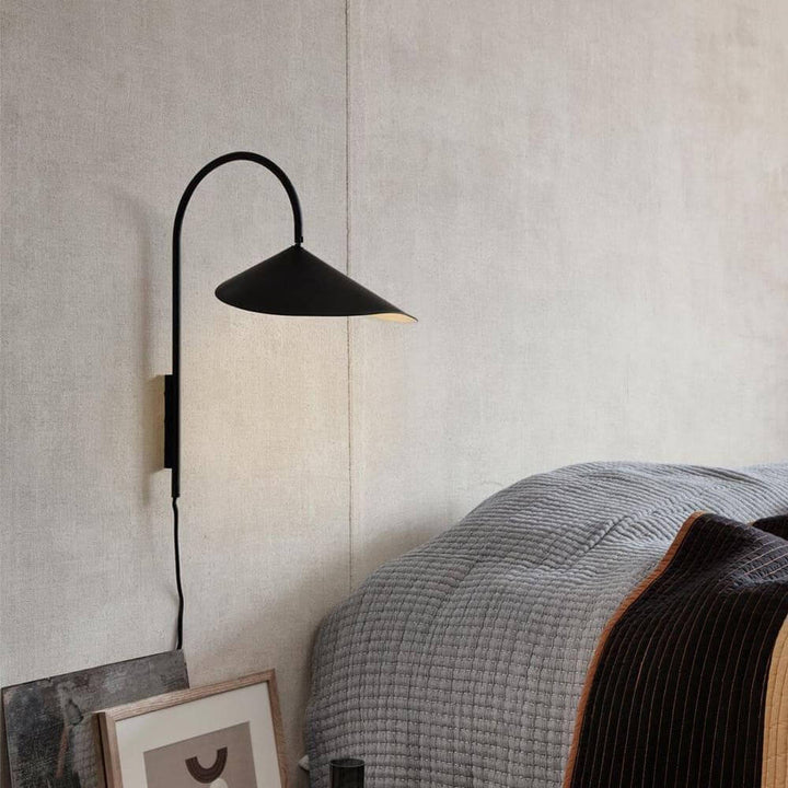 Unlit black Arum plug-in wall sconce mounted beside a gray upholstered bed