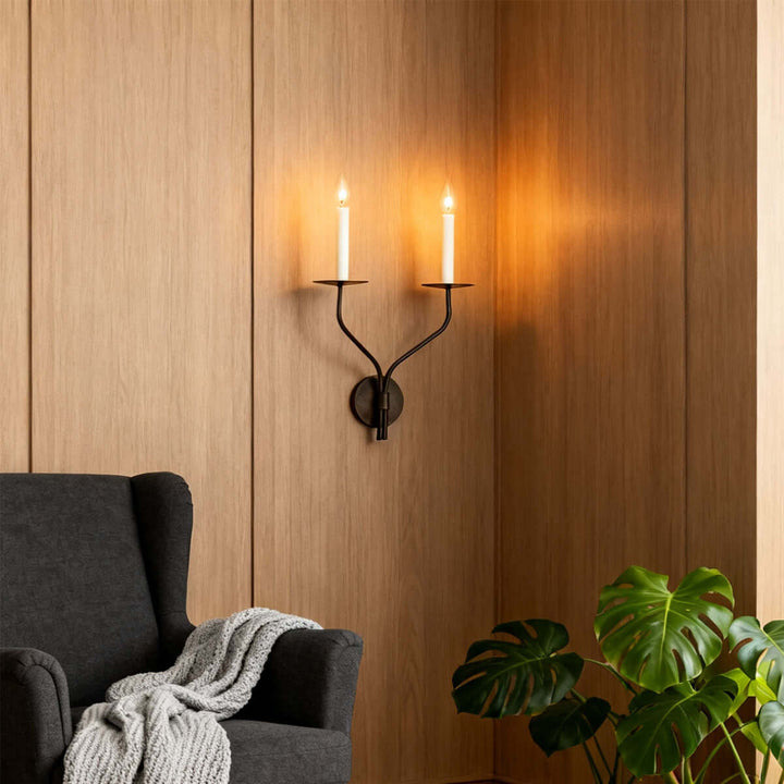 Black two-light candle wall sconce mounted on wood paneling beside a living room armchair