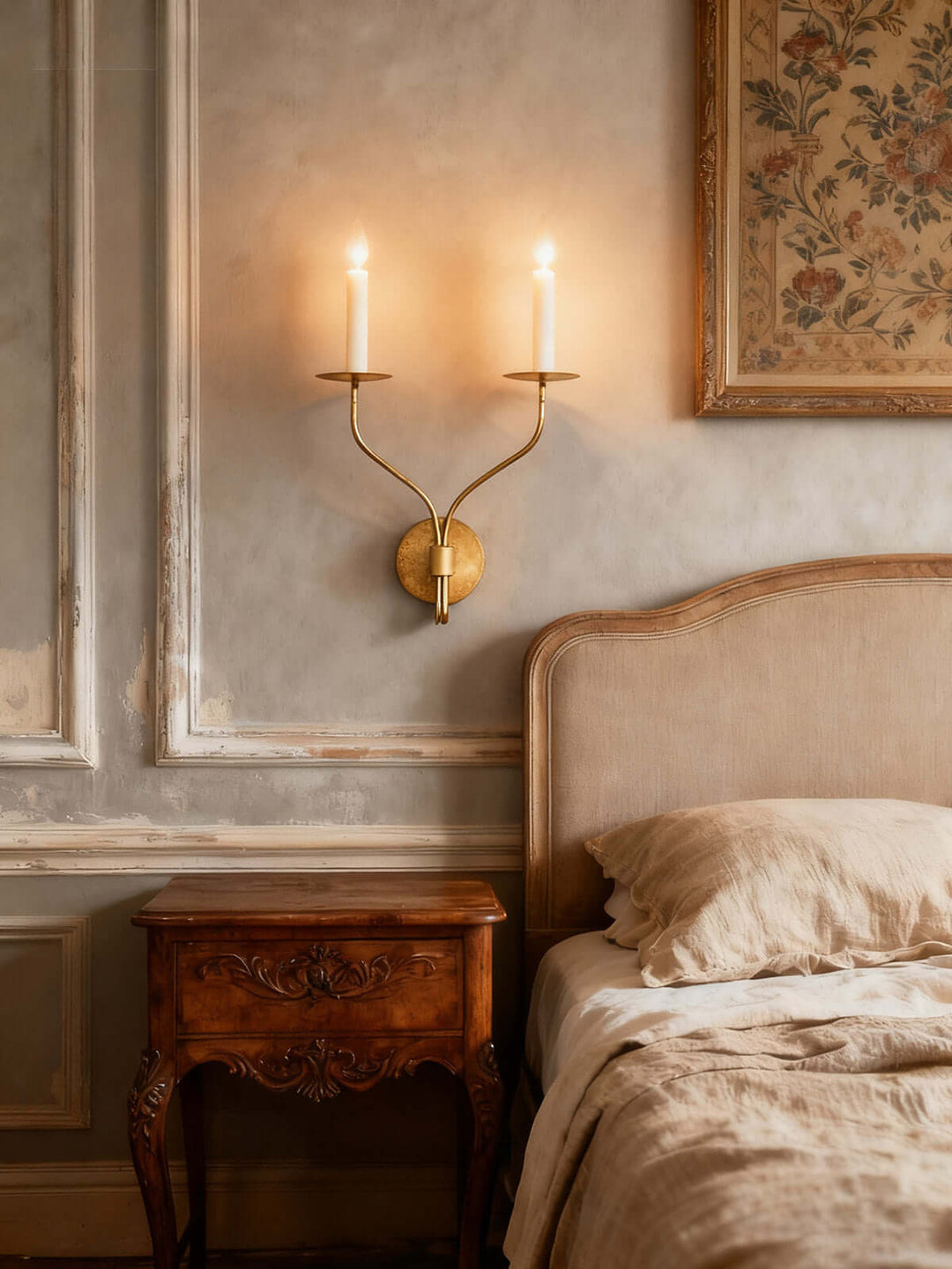 Aged brass two-light candle wall sconce casting warm light above an upholstered bedroom headboard