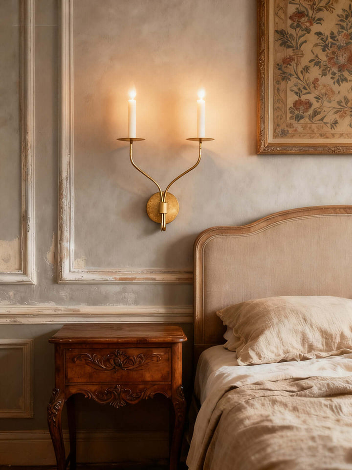 Aged brass two-light candle wall sconce casting warm light above an upholstered bedroom headboard