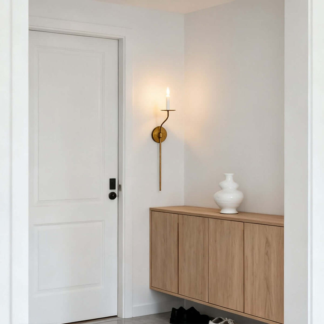 Tall brass candle wall sconce providing warm entryway lighting beside a wood storage cabinet