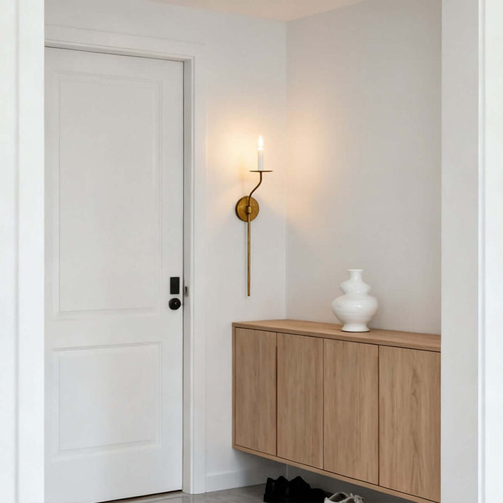 Tall brass candle wall sconce providing warm entryway lighting beside a wood storage cabinet
