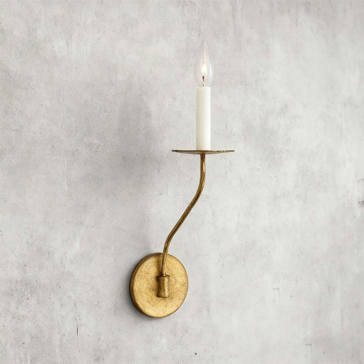 Short aged brass single-light candle wall sconce with round backplate on a textured gray wall