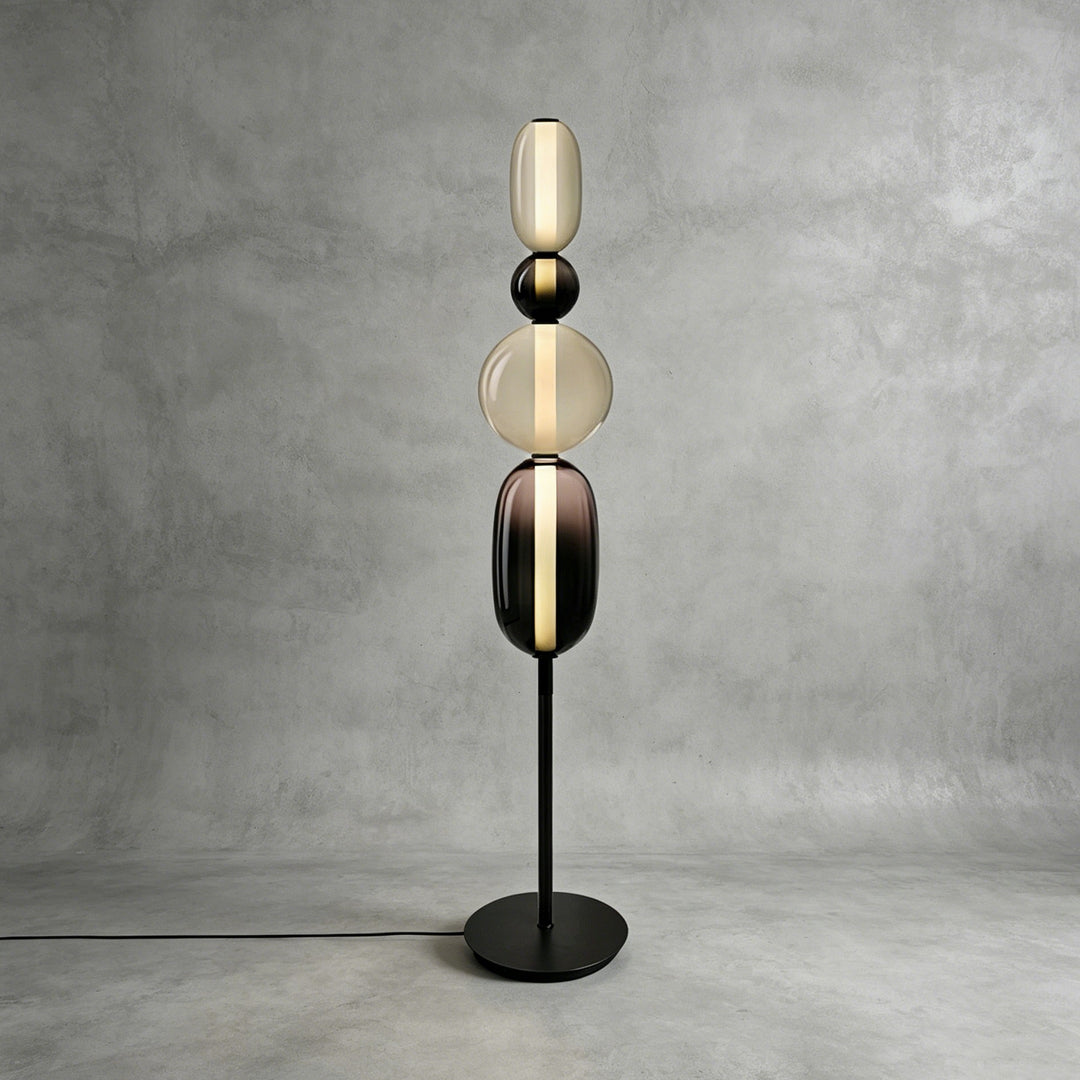 Tall Candied Model B Black glass floor lamp glowing against a textured gray studio wall.