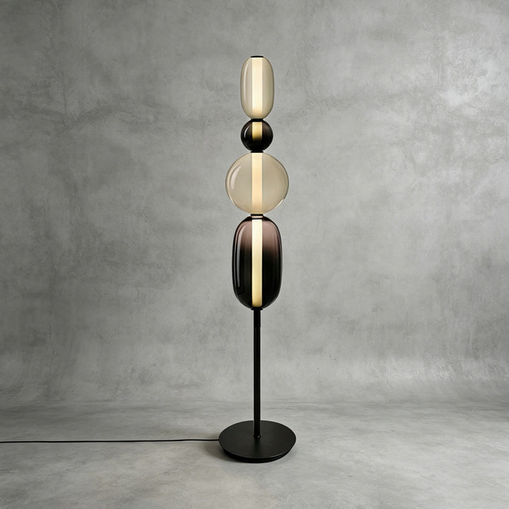 Tall Candied Model B Black glass floor lamp glowing against a textured gray studio wall.