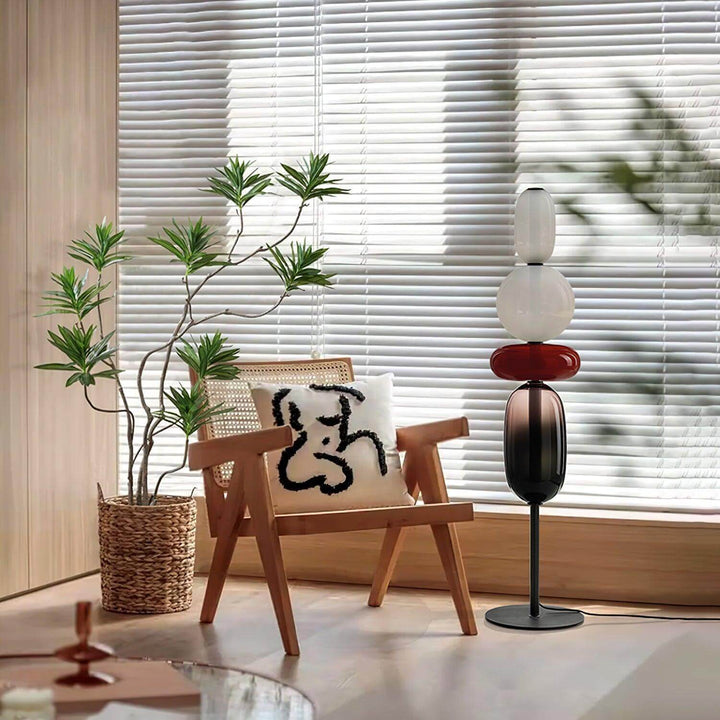 Unlit Candied red glass floor lamp beside a wood lounge chair, potted plant, and window blinds.