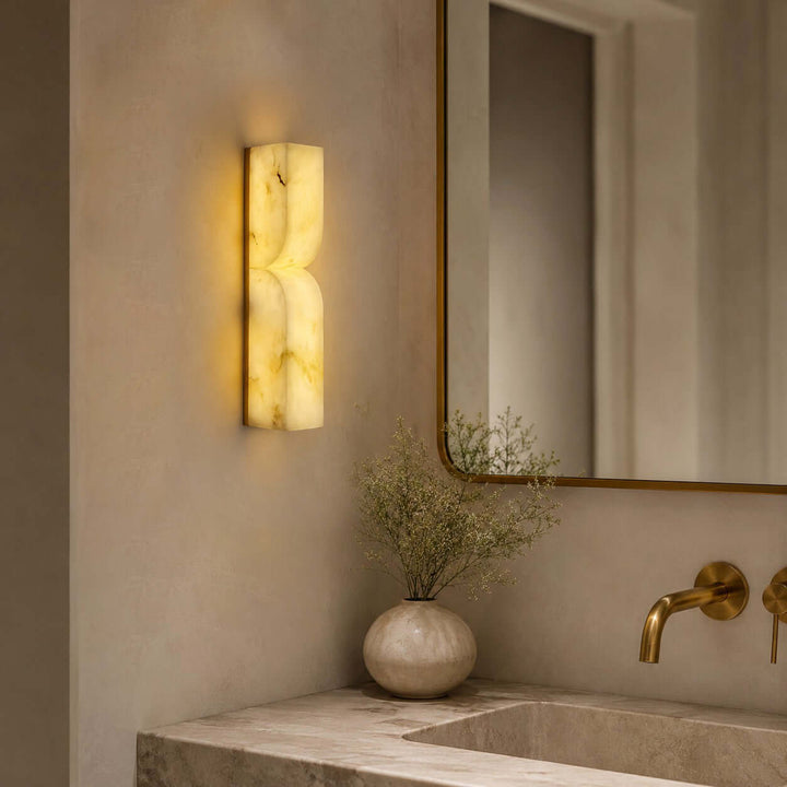 Cavira alabaster wall sconce glowing beside a brass-framed mirror and stone vanity in a modern bathroom