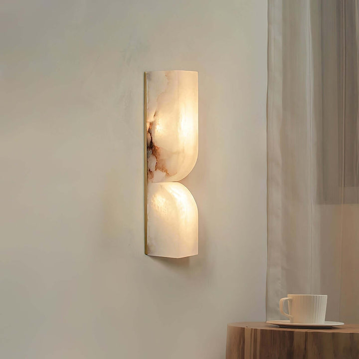 Cavira alabaster wall sconce casting soft light beside a sheer curtain and small wood accent table