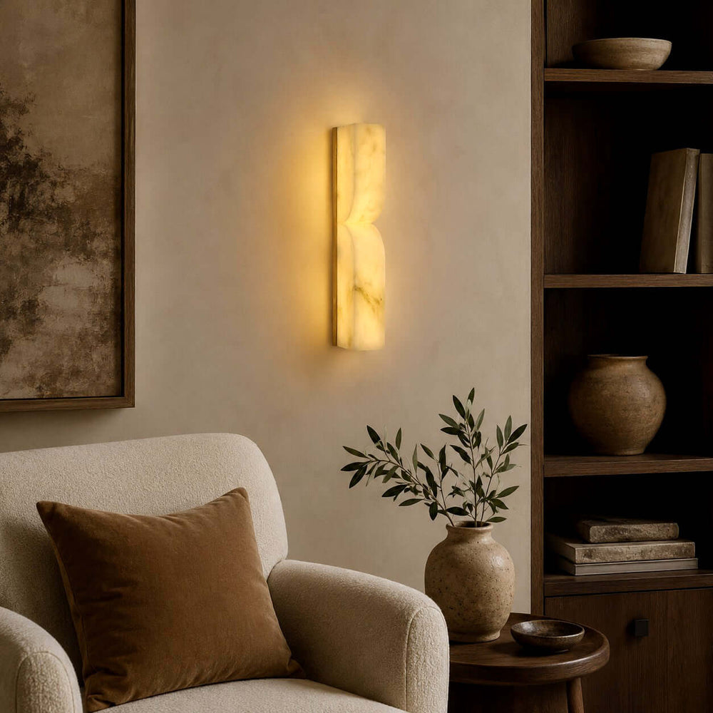 Cavira alabaster wall sconce casting a warm glow beside an upholstered chair in a neutral living room