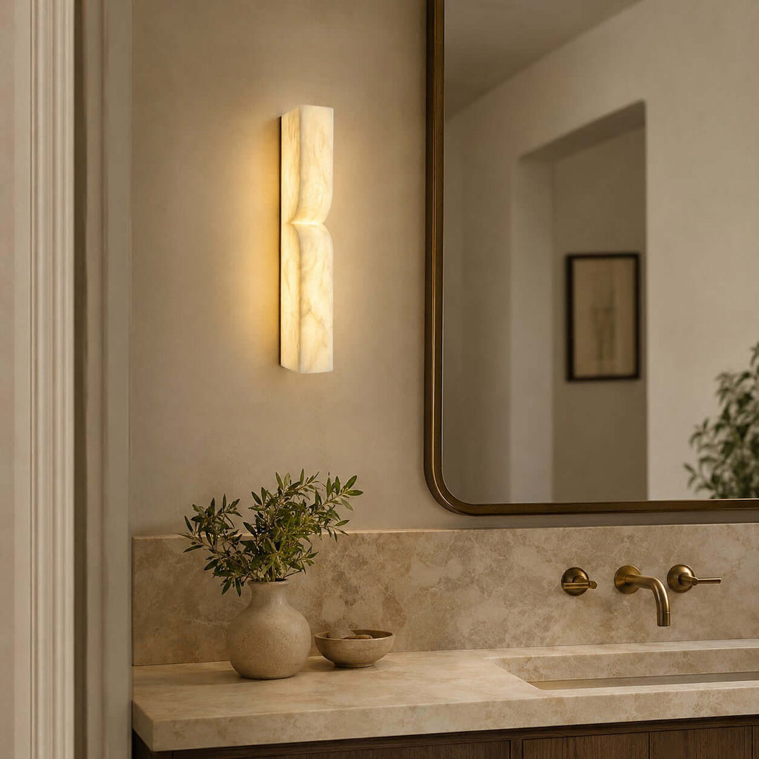 Cavira wall sconce beside a brass-framed bathroom mirror and stone vanity with a soft diffused glow