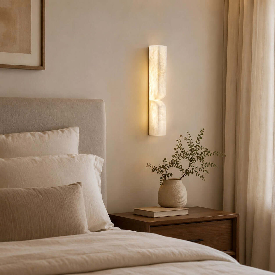 Cavira wall sconce casting soft light beside an upholstered bed and wood nightstand in a neutral bedroom
