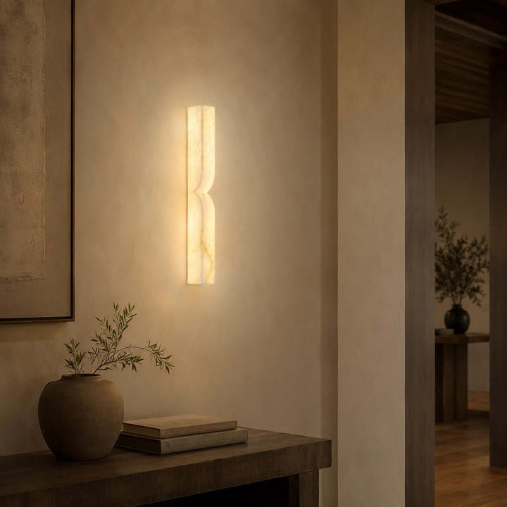 Cavira alabaster wall sconce creating a warm glow above a rustic wood console in a quiet hallway