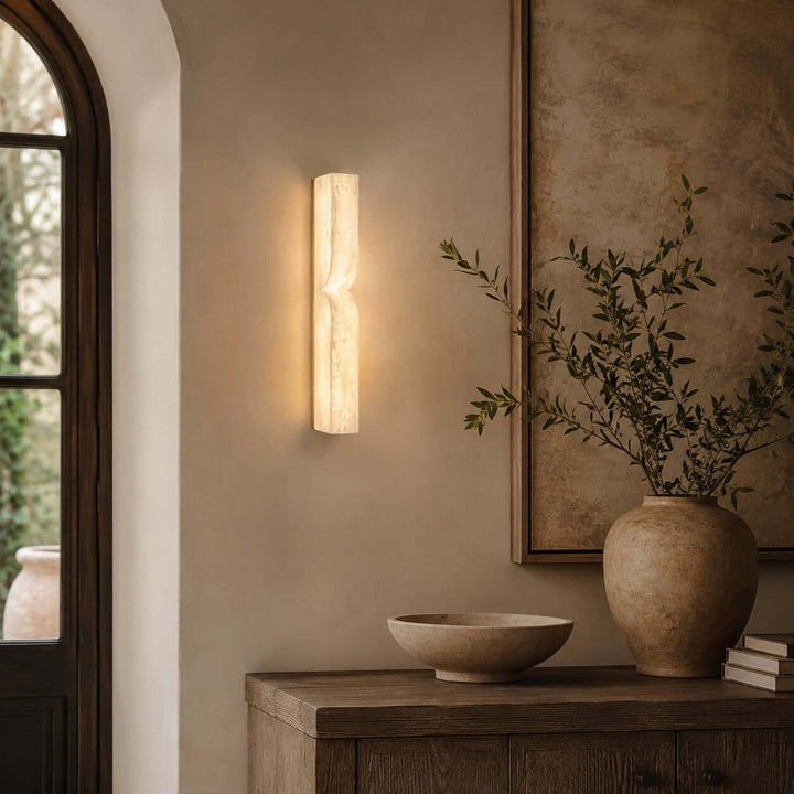 Cavira alabaster wall sconce glowing above a wood sideboard beside an arched glass entry door