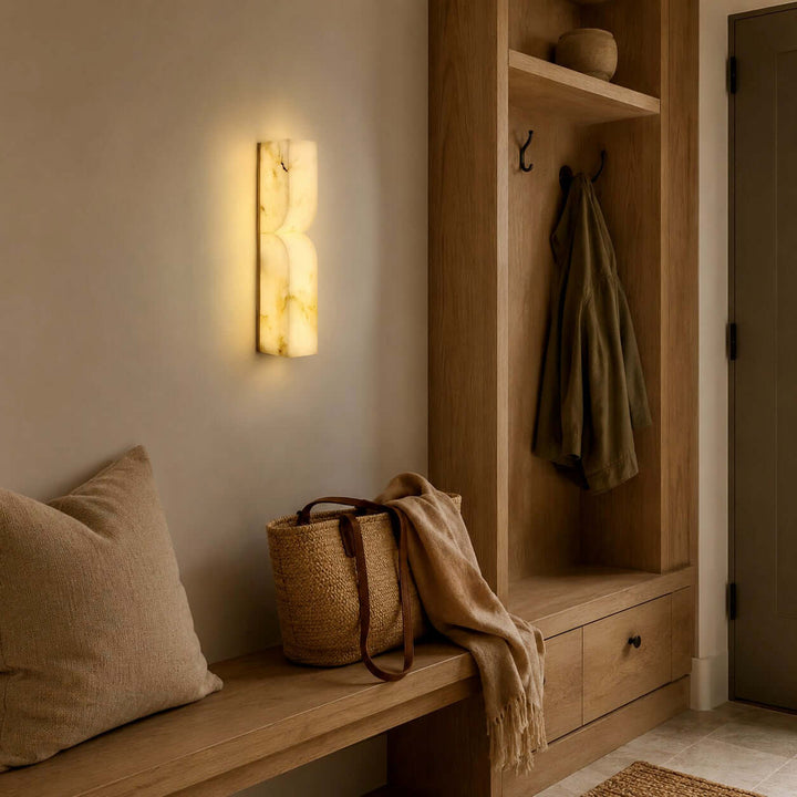 Cavira alabaster wall sconce lighting a warm mudroom entry with a wood bench, coat hooks and woven basket