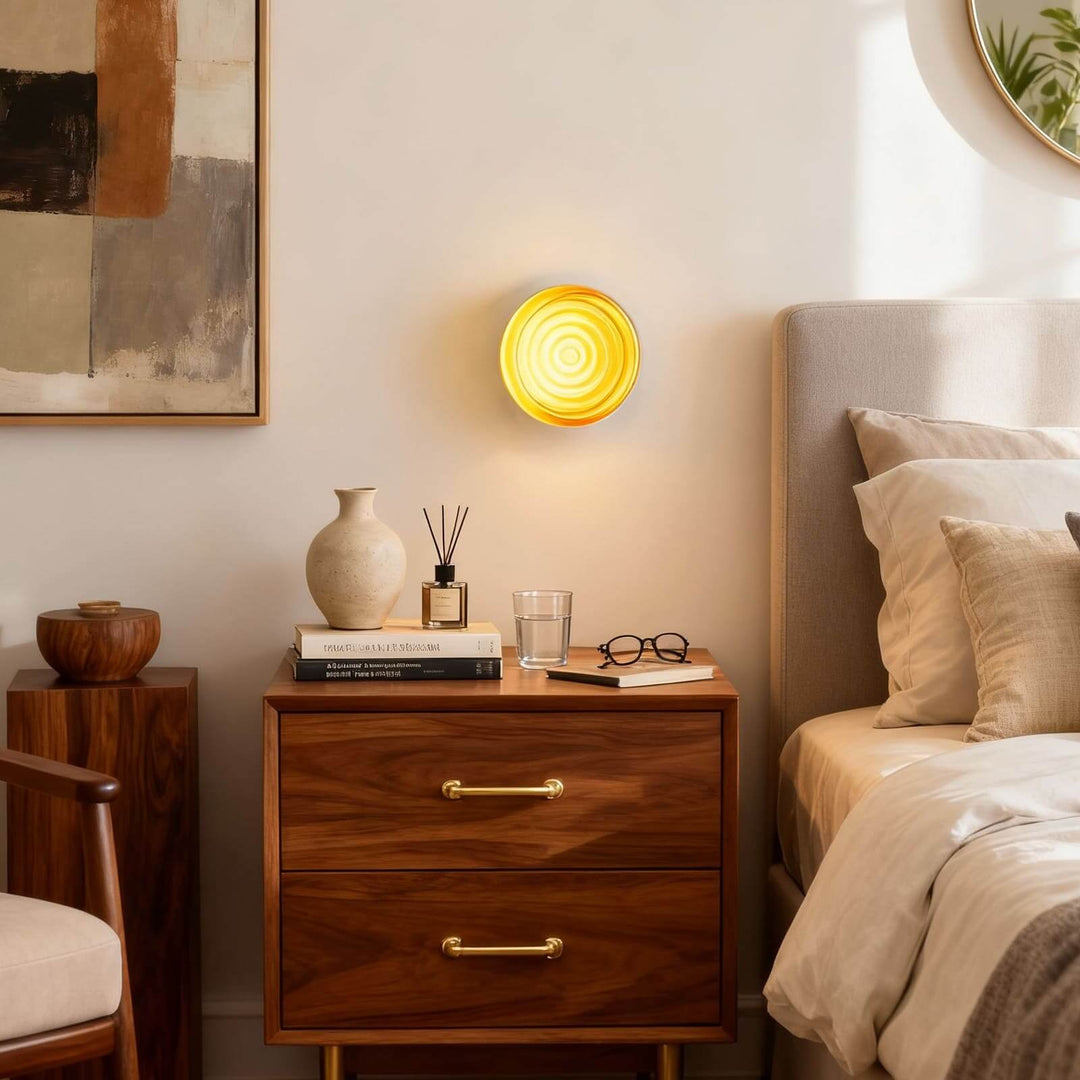 Amber round wall sconce above a wood nightstand with books and bedside decor.