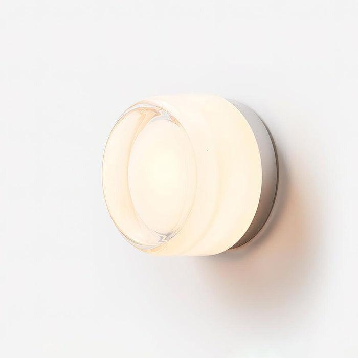 White glass wall sconce lit on a plain background with a clear front lens and metal backplate.