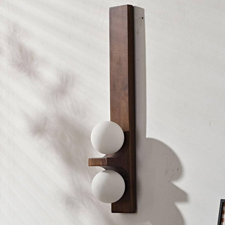 Tall dark wood double wall sconce with two white glass globes shown unlit on a white wall