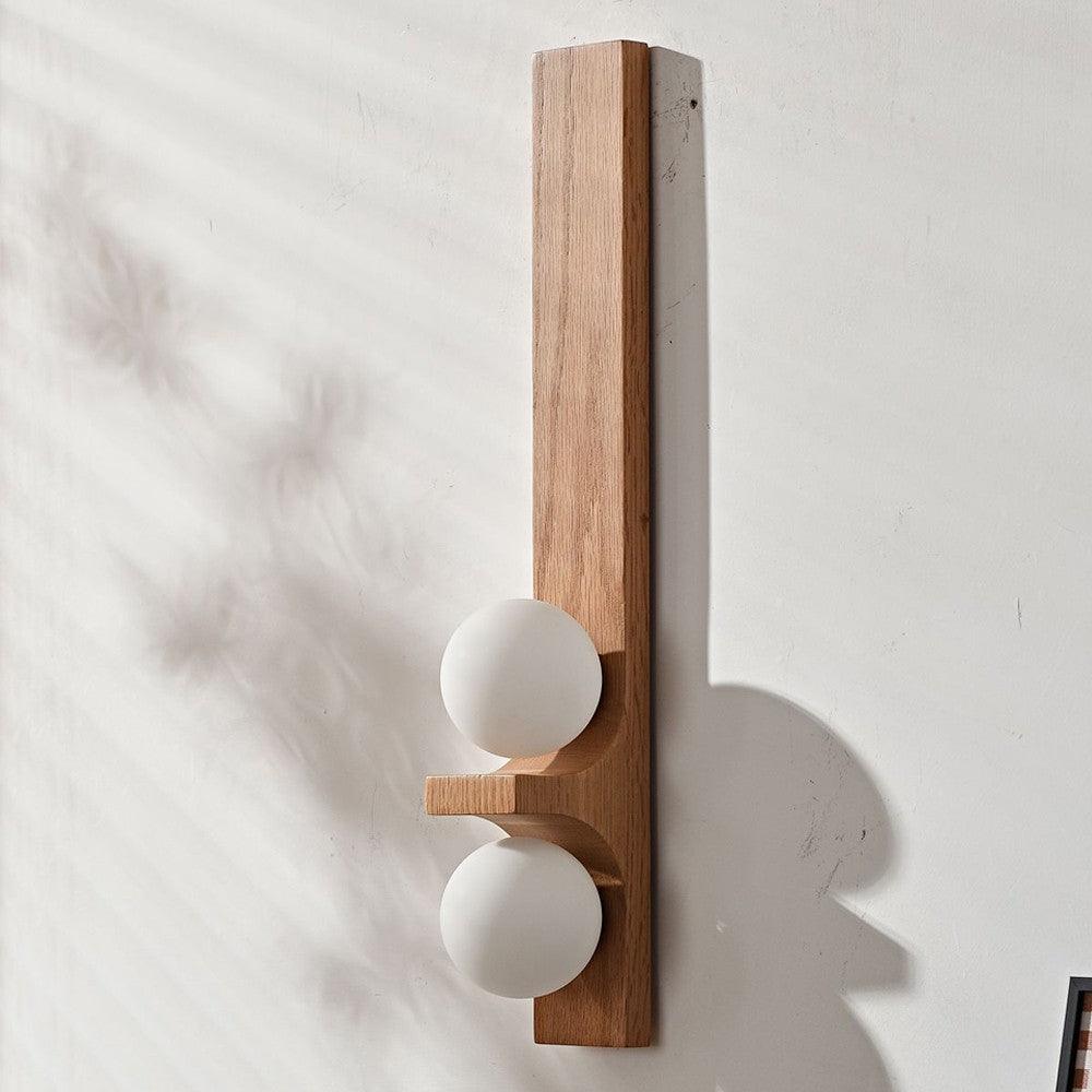 Tall light wood double wall sconce with two white glass globes shown unlit on a white wall