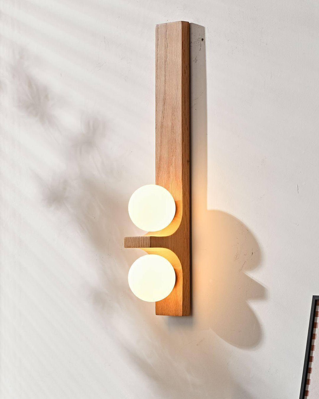 Tall light wood double wall sconce with two glowing white globes casting warm light on a wall