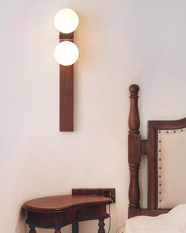 Tall wood double wall sconce lit above a vintage bedside table and upholstered bed frame