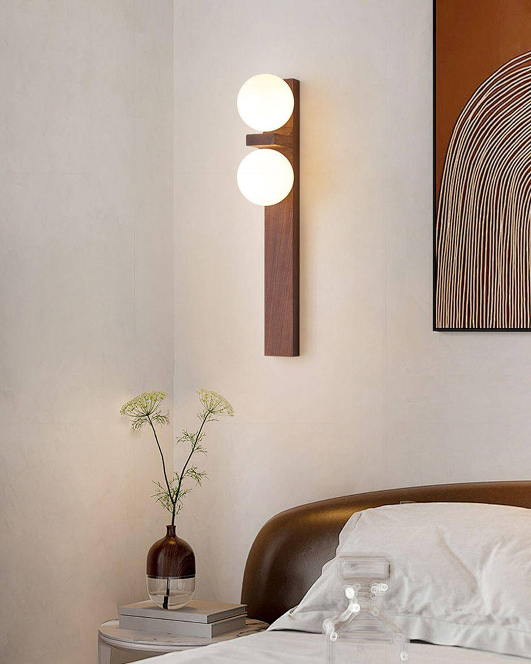 Tall wood double wall sconce glowing above a bedside table with books vase and wall art