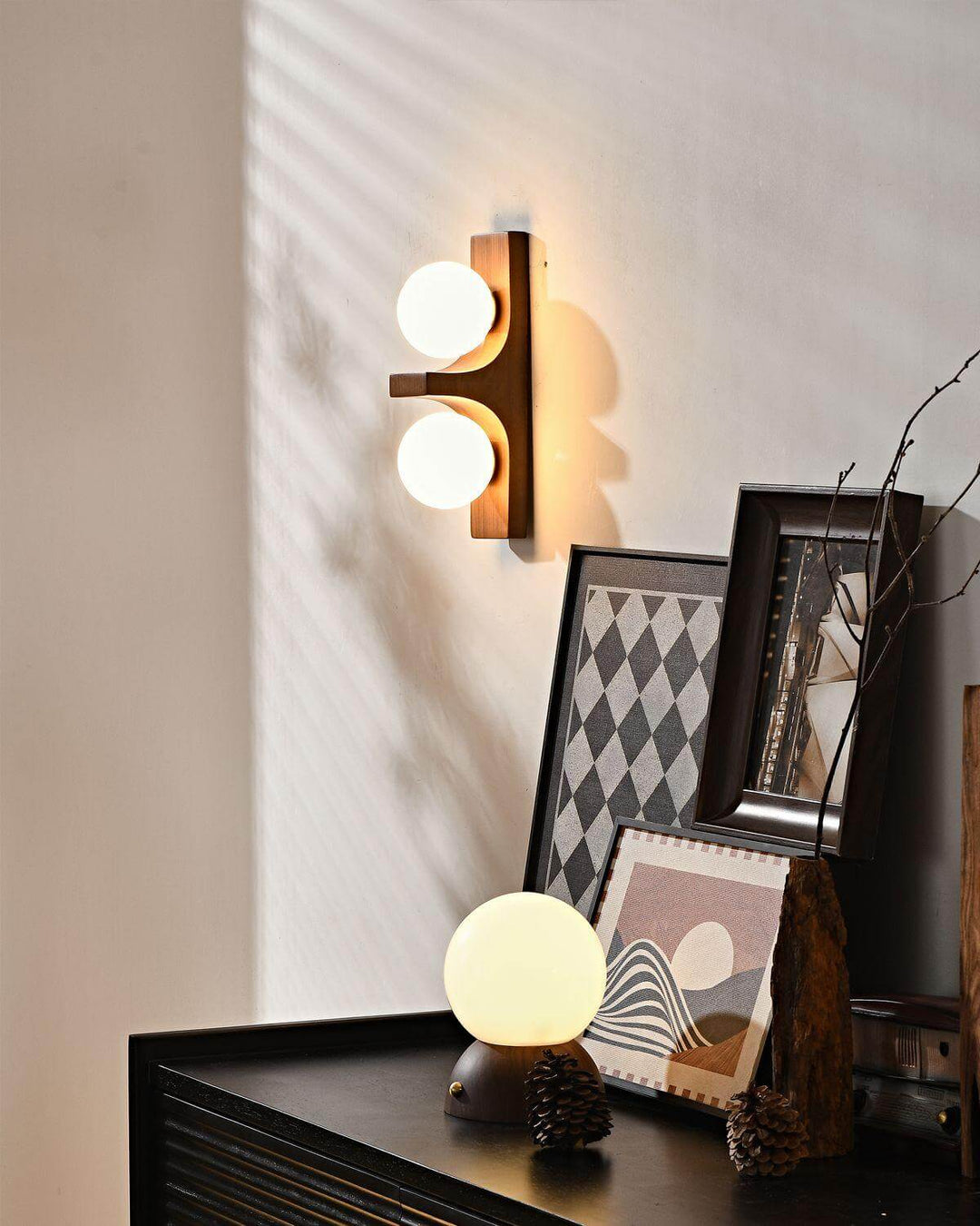 Short wood double wall sconce lit above a black cabinet with framed art and a glowing table lamp