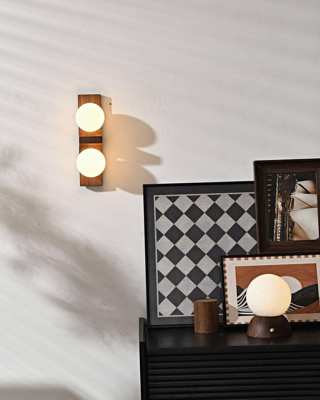 Short wood double wall sconce glowing above a black cabinet with framed art and a table lamp